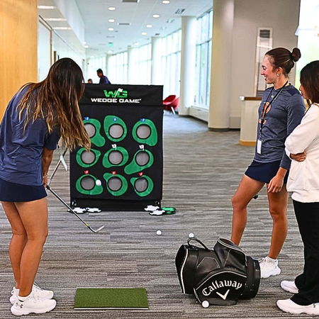 Three women practicing golf swings in a room with a wedge game target and Callaway bag.